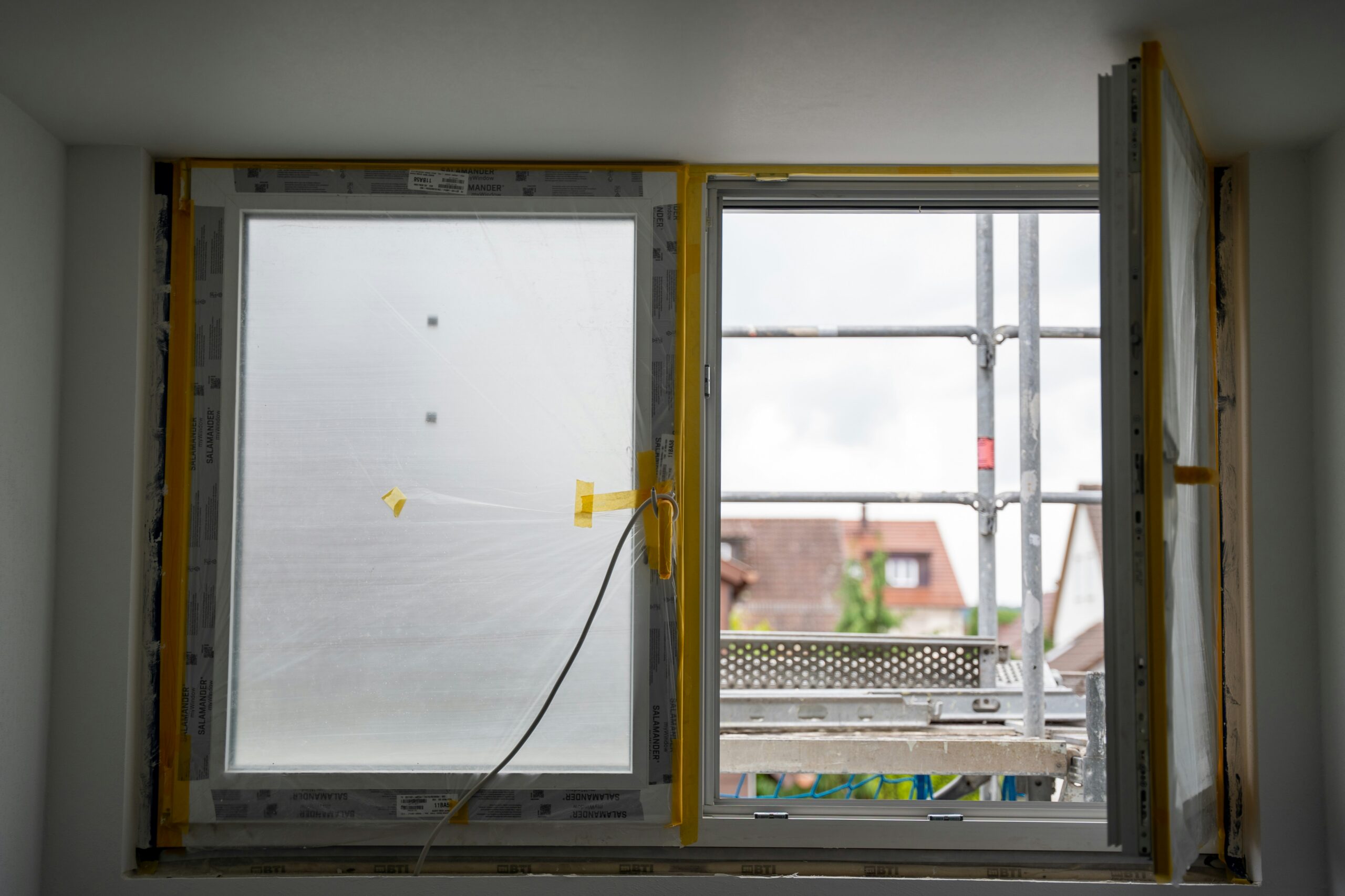 A worker fits a new white-framed window into the exterior wall of a residential home on a sunny day.