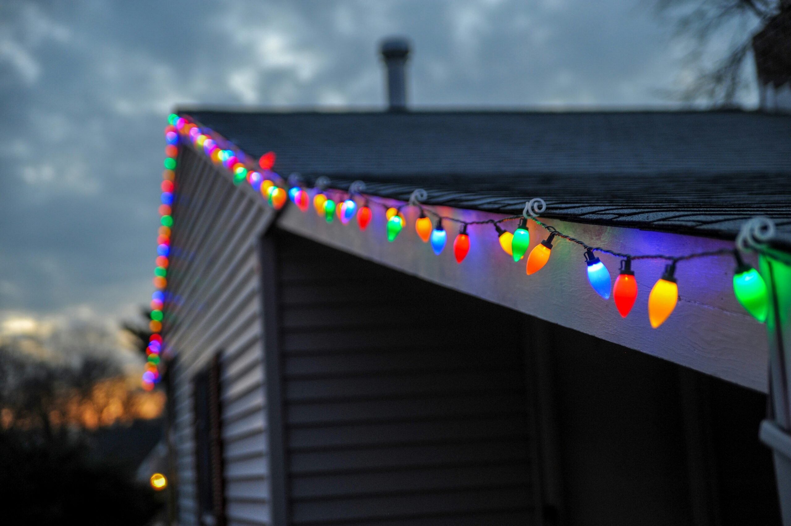 Strand of holiday lights attached with plastic clips along the gutter of a house with a shingled roof.